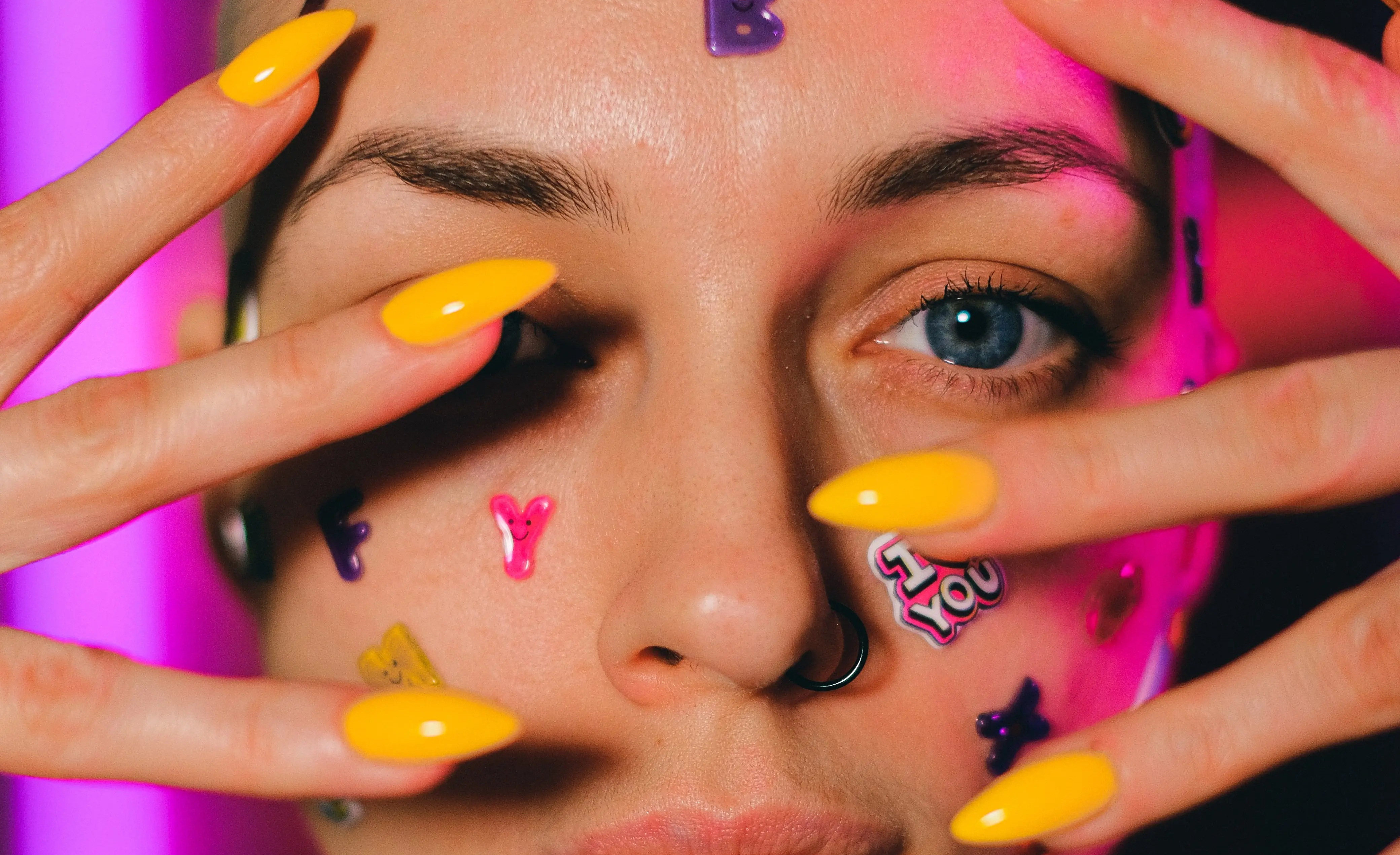 Close-up photo of a white woman with a shaved head and bright yellow nails, with stickers all over her face
